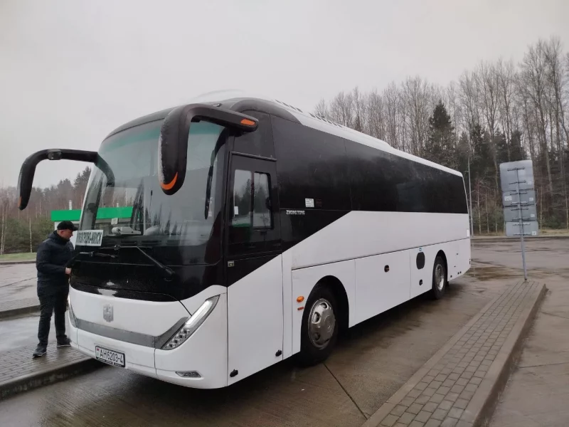 На рэйсах Гродна-Мінск замест маршрутак з'явіліся вялікія аўтобусы Вялікі белы аўтобус Zhong Tong, прыпаркаваны на мокрым тратуары каля дрэў, з чалавекам, які стаіць побач, адкідвае адзінокую постаць на фоне хмарнага неба трасы Гродна-Мінск.